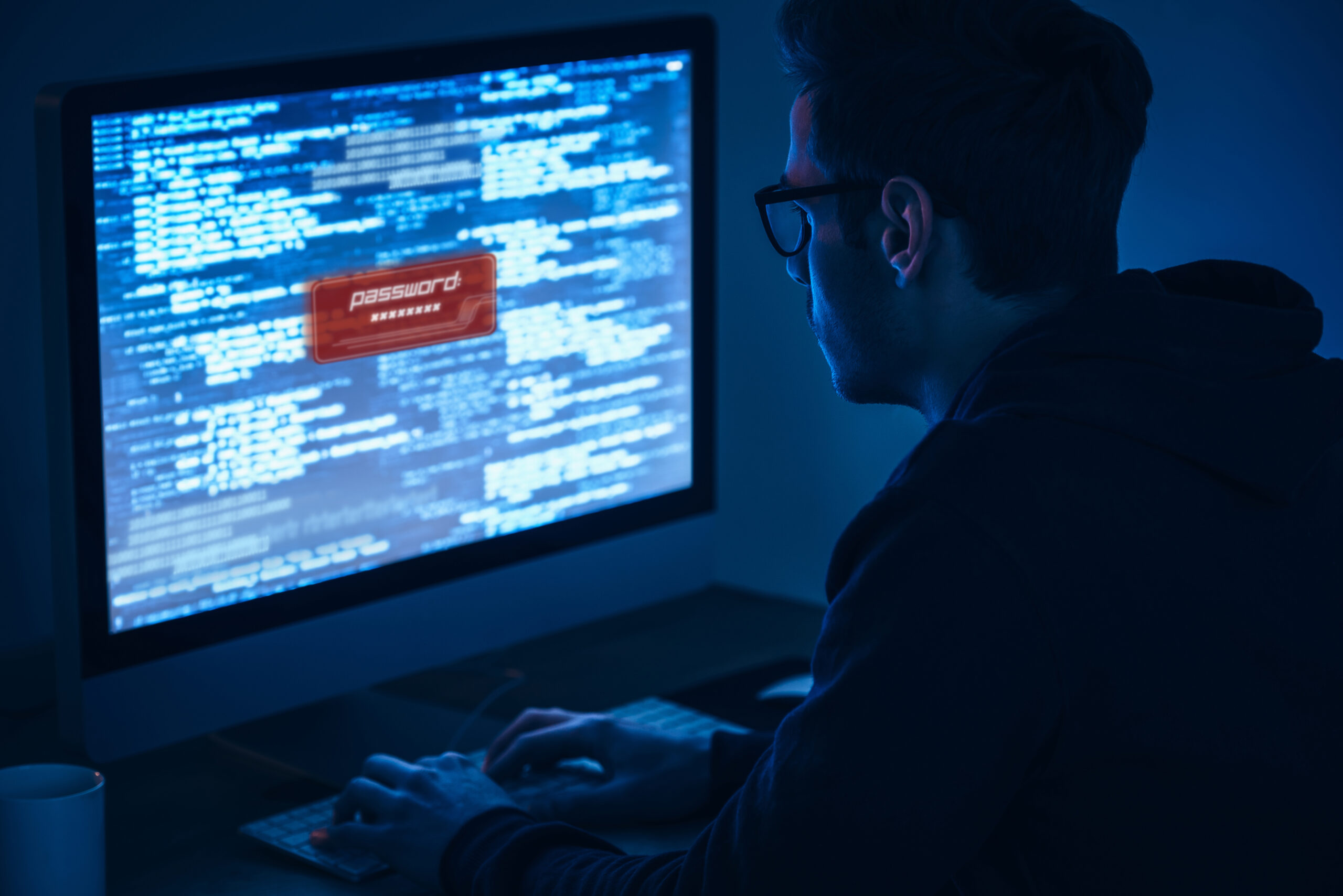 Computer security. Rear view of young man typing and looking at computer monitor while sitting at the table in dark room